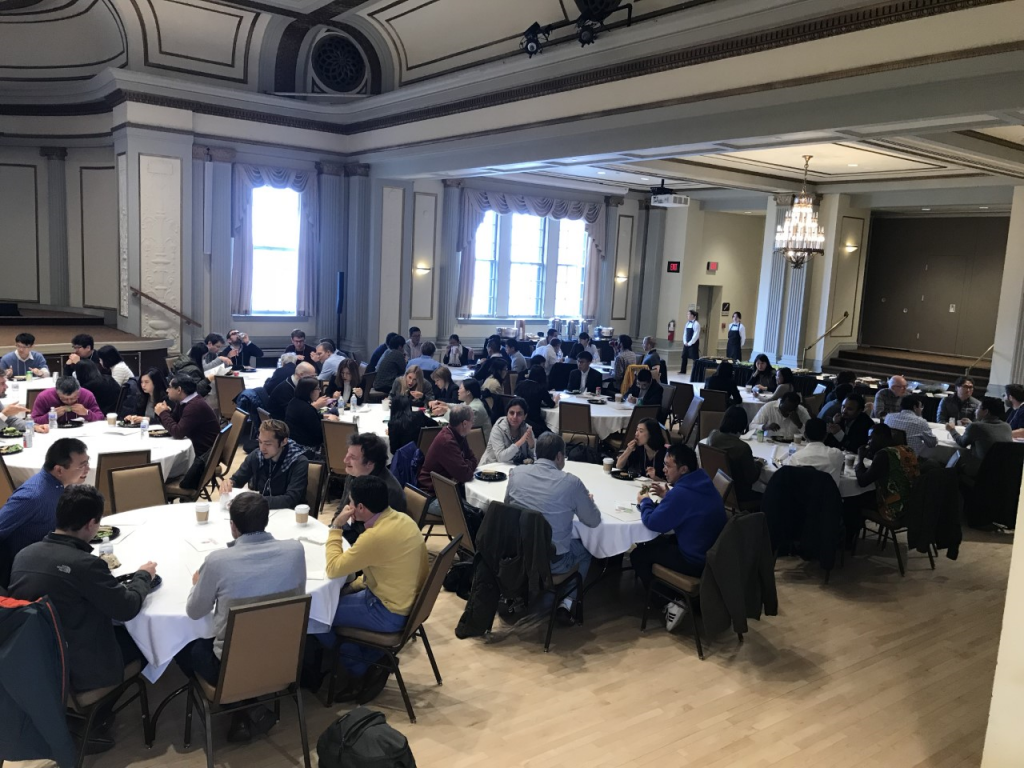 people sit at tables, eat and talk before a presentation starts