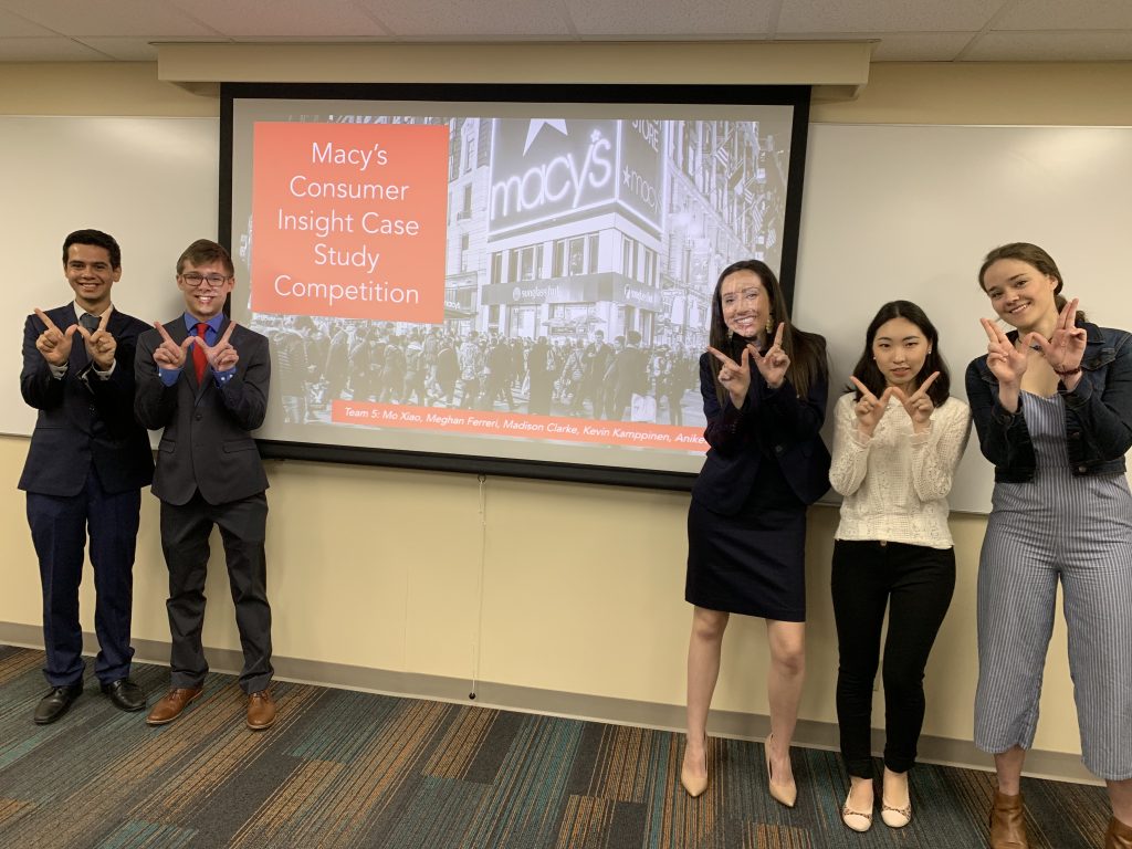 Students stand in front of a screen before their presentation about Macy's consumer insights for the undergraduate case competition holding up a 'W' with their hands