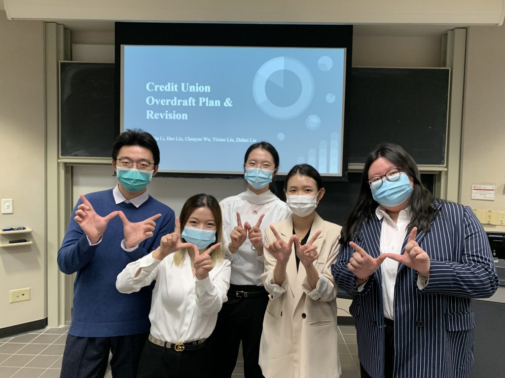 Students stand in front of a screen before their presentation about the Credit Union Overdraft Plan & Revision while holding up a 'W' with their hands