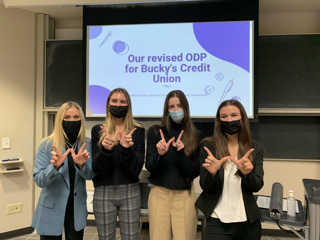 Students stand in front of a screen before their presentation about their revised ODP for Bucky's Credit Union while holding up a 'W' with their hands