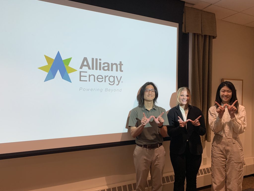 Three Students pose in front of a projector with the Alliant Energy logo behind them before their Case Competition presentation