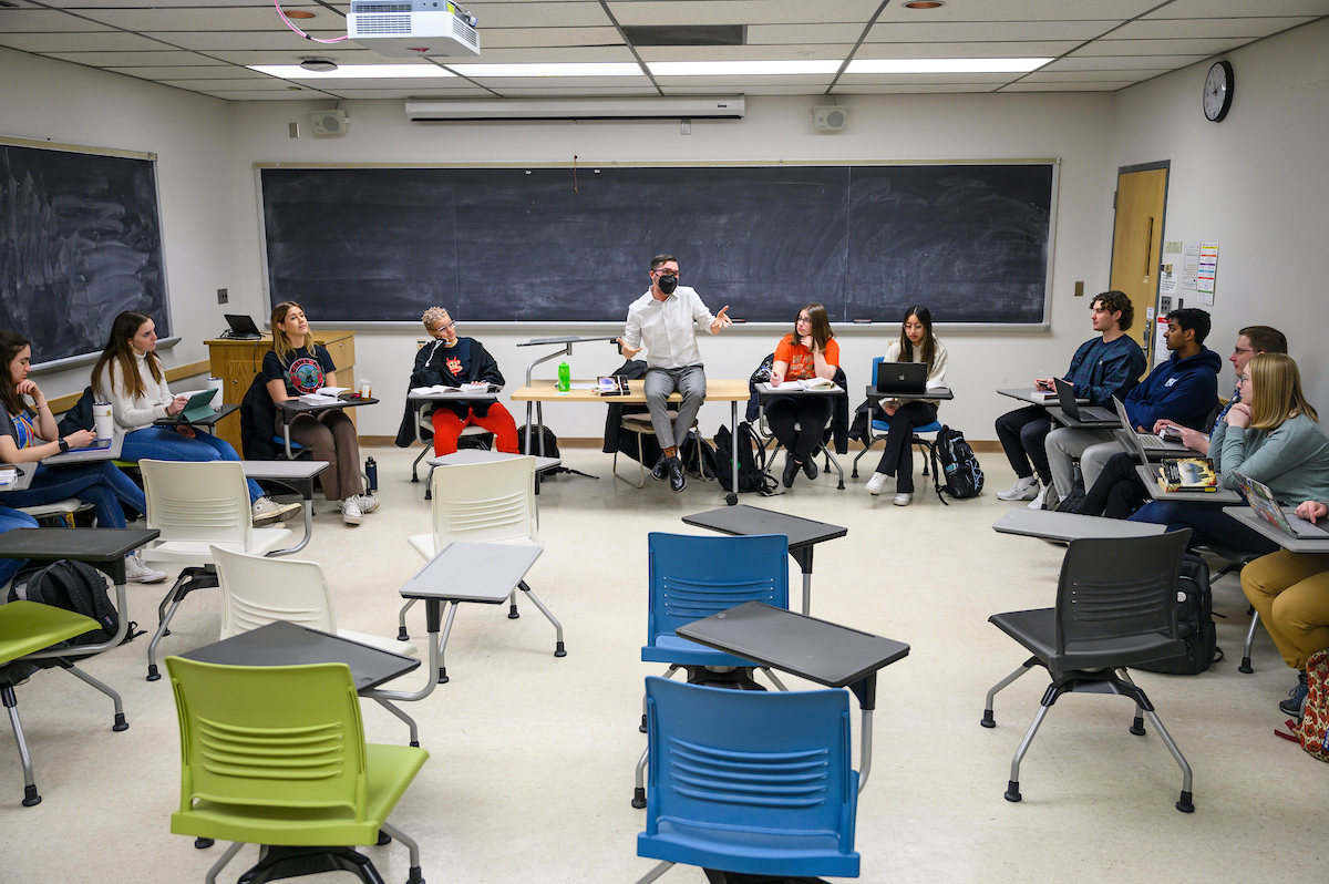 Students sit in a circle in their classroom and listen to their professor speak.